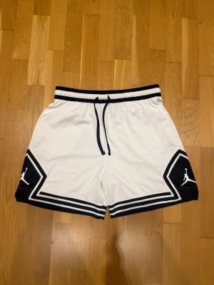 Jordan shorts mesh - Snygga vita basketshorts från Jordan med svarta kontrastdetaljer och logga på benen. Shortsen har snörning i midjan och är tillverkade i ett ventilerande polyestermaterial. Perfekta för sport eller en avslappnad stil.                                                           Modellen är 185cm lång och väger 82kg.