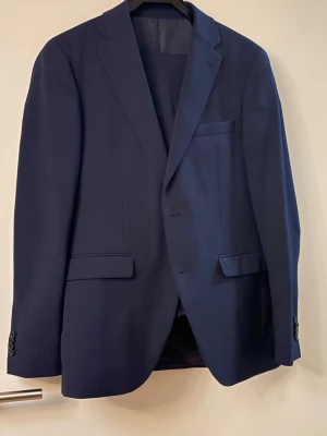 Mörkblå kostym  från Volt från Riley Stanton - Snygg mörkblå kostym från Riley, modell Stanton. Klassisk design med två knappar framtill, fickor med lock och bröstficka. Fodrad insida med elegant finish. Perfekt för dig som vill ha en stilren och tidlös look. Perfekt för kommande bal eller student eller bröllop 