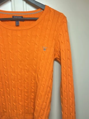 Orange kabelstickad tröja från Gant - Snygg orange kabelstickad tröja från Gant med rund halsringning och diskret logga broderad på bröstet. Tröjan har långa ärmar och ribbade muddar vid ärmslut och nederkant. Perfekt för dig som vill ha en färgstark och klassisk look.