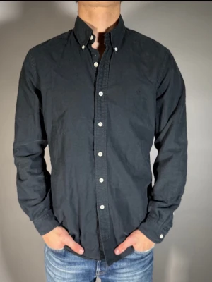 Svart skjorta från Ralph Lauren - Klassisk svart skjorta från Ralph Lauren med button-down krage och vita knappar. Skjortan har lång ärm och diskret broderad logga på bröstet. Tillverkad i mjuk bomull och passar perfekt till jeans eller chinos.