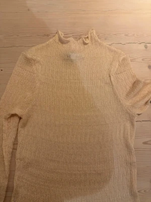 Beige ribbad polotopp - Trendig beige polotopp med ribbad struktur och lång ärm. Toppen har en hög krage och är tillverkad i ett mjukt, stretchigt material som ger en snygg passform. Perfekt att styla med jeans eller kjol för en clean look.