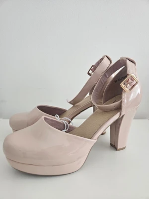 Nude lackpumps med ankelrem från VOX - Snygga pumps i nude färg från VOX med glansig lackyta och bred klack. Skorna har rund tå, platåsula och justerbar ankelrem med guldfärgat spänne. Veganskt material och stilren design som passar till många outfits.