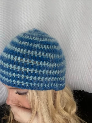Blå- och ljusblå randig stickad mössa - Säljer en handvirkad mössa i blå och ljusblå ränder. Mössan är virkad i ett fluffigt och mjukt garn. Den har en klassisk form som sitter tajt på huvudet och passar perfekt för kalla dagar.