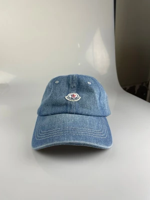 Moncler denimkeps med broderad logga - Säljer en snygg denimkeps från Moncler i ljusblå jeans med broderad logotyp framtill. Klassisk böjd skärm och justerbar baksida. Perfekt för dig som vill ha en stilren och trendig accessoar till vardagslooken.