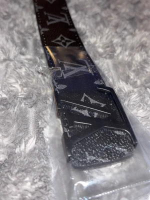 Svart L bälte med monogram - Snyggt svart bälte från L med klassiskt L monogram i silverfärg över hela bandet. Bältet har en matchande L spänne i metall och är gjort i präglat skinn. Perfekt accessoar för att lyfta vilken outfit som helst.