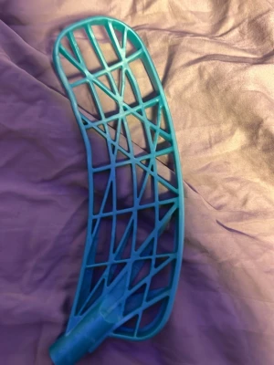 Blått innebandyblad i plast - Säljer ett blått innebandyblad med ett öppet rutmönster och böjd form. Bladet är gjort av plast och har ett fäste för skaft. Perfekt för dig som vill byta blad på din klubba eller testa något nytt på planen.