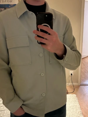 Ljusgrön overshirt från Zara - Snygg ljusgrön overshirt från Zara med raka linjer och stora bröstfickor. Klassisk krage och knäppning framtill med matchande knappar. Tillverkad i ett mjukt tyg som känns bekvämt och stilrent. Perfekt lager på lager-plagg för en clean look.