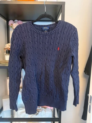 Mörkblå kabelstickad tröja Polo Ralph Lauren - Kabelstickad mörkblå tröja från Polo Ralph Lauren med rund halsringning och den klassiska röda logotypen broderad på bröstet.  Rätt så urtvättad men annars fin❤️