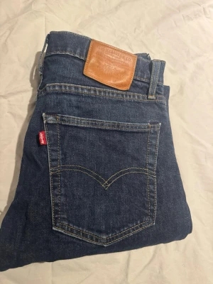 Levi's 502 mörkblå jeans W30 L32 - Klassiska Levi's 502 jeans i mörkblå tvätt med raka ben och fem fickor. Jeansen har den ikoniska röda Levi's-taggen på bakfickan och brunt läderpatch i midjan. Tillverkade i slitstarkt bomullsjeansmaterial och har knappgylf framtill.