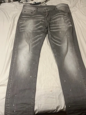 Grå Dsquared2 jeans med slitningar - Säljer ett par grå jeans från Dsquared2 med coola slitningar och vita färgstänk. Jeansen har klassisk femficksmodell, raka ben och detaljerade sömmar. Materialet är denim med en snygg tvättad look och unika effekter. Perfekta för dig som gillar streetstyle och statement-plagg.