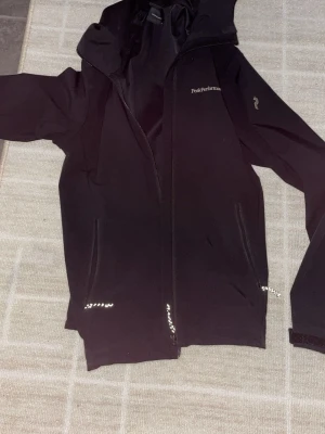 Svart vindjacka från Peak Performance - Svart vindjacka från Peak Performance med huva och dragkedja framtill. Två sidofickor med dragkedja och diskret logga på bröstet samt ärmen. Tillverkad i ett lätt och smidigt material som passar perfekt för blåsiga dagar.