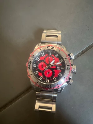 Festina kronografklocka i silver och rött - Snygg kronografklocka från Festina med robust länkarmband i silverfärgat metall. Urtavlan är svart med röda detaljer och flera subdials, samt datumvisning. Klockan har markerade siffror runt boetten och tydliga knappar på sidan för funktioner.