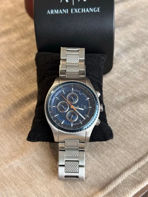 Armani Exchange silverklocka blå urtavla - Stilren klocka från Armani Exchange med silverfärgad länk i metall och blå urtavla. Klockan har tre mindre urtavlor, kronograf och tydliga indexmarkeringar. Snygg detalj med texturerade länkar och logga på spännet. Perfekt accessoar för dig som gillar klassisk men modern stil. Extra länkar följer med.