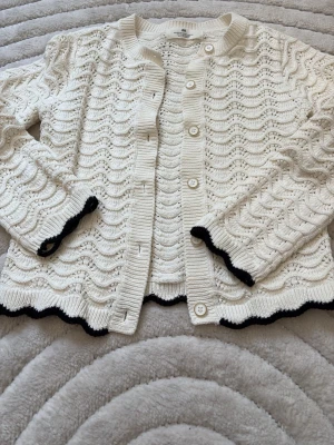 Vit virkad kofta med svarta detaljer - Snygg vit kofta i virkad look med vågigt mönster och svarta kanter på ärmslut och nederkant. Koftan har rund hals, knappar framtill och lång ärm. Perfekt för dig som vill ha lite blair waldorf vibes.