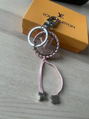 Vuitton nyckelring med rosa detaljer - Säljer en exklusiv Vuitton nyckelring med silverfärgade metalldetaljer, rosa flätad kant och två rosa band med blommor i metall. Perfekt som accessoar till väskan eller nycklarna. Kommer i originalförpackning.