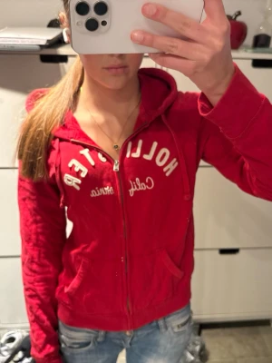 Hollister röd zip hoodie  - Röd zip up hoodie från Hollister med dragkedja och vit broderad text på bröstet.                                     Storlek L men passar S bättre!                          Hoodien har huva med dragsko och två fickor framtill. Perfekt för en avslappnad och cool stil. 