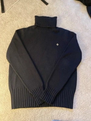 Mörkblå polotröja med brodyr - Mysig mörkblå stickad polotröja med ribbade muddar och hög krage. Tröjan har en liten broderad logga på bröstet och är tillverkad i ett tjockare material som passar perfekt när det är kallt ute.