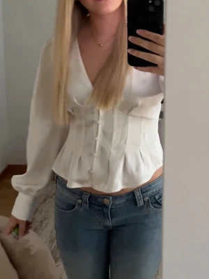 Vit satinblus med knappar och v-ringning - Stilren vit blus från vero Moda i glansigt satinmaterial. Blusen har v-ringning, markerad midja och dekorativa knappar framtill. Långa puffärmar med breda manschetter ger en trendig siluett. Sparsamt använd och är som i nyskick! Storlek M🩷