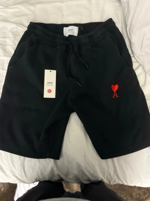 Svarta shorts från Ami med rött hjärta - Svarta shorts från Ami med dragsko i midjan och två sidofickor. På vänster ben finns ett broderat rött hjärta med bokstaven A. Sköna och stilrena, perfekta för sommaren. Materialet känns mjukt och är troligtvis bomull.
