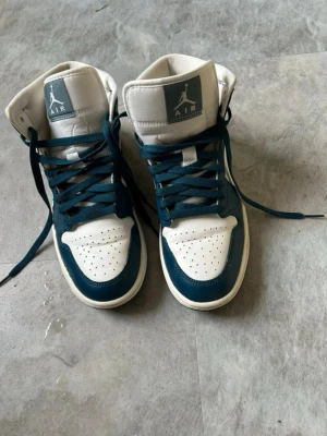 Air Jordan 1 Mid blå/vit sneakers - Säljer ett par Nike Air Jordan 1 Mid sneakers i blått och vitt. Skorna har högt skaft, klassisk snörning och den ikoniska swoosh-loggan på sidan. Perfekta för dig som gillar streetwear och vill ha en stilren look. Dessa är använda 3 gånger sedan endast stått hemma, så är i otroligt bra skick! Storlek 40.