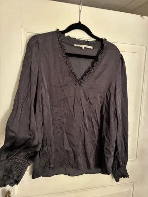 Mörkgrå blus med volangkant Second Female - Snygg mörkgrå blus med v-ringning och volangkant runt halsen från Second Female. Blusen har långa ärmar med smock vid ärmslut och en avslappnad passform. Perfekt att styla med jeans eller kjol för en trendig look.