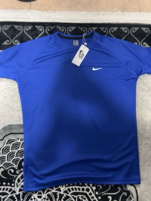 Blå Nike t-shirt i funktionsmaterial - Snygg blå t-shirt från Nike med klassisk vit logga på bröstet. Tillverkad i ett lätt och ventilerande funktionsmaterial som passar perfekt till träning. T-shirten har rund halsringning och korta ärmar för en sportig look.