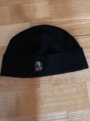 Svart mössa från Parajumpers - Säljer en svart ribbstickad mössa från Parajumpers med uppvikt kant och klassisk logopatch framtill. Perfekt för kalla dagar och enkel att matcha med olika outfits. Materialet känns mjukt och bekvämt mot huden.