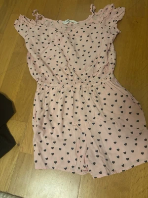 Rosa kort klänning med hjärtan - Supersöt rosa kort klänning med svarta hjärtan och resår i midjan. Klänningen har volangdetaljer vid axlarna och är ärmlös, perfekt för varma dagar. Mjuk och skön känsla, troligen i bomull eller polyester.