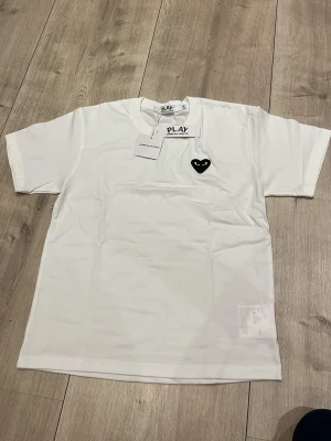 Vit Comme des Garçons PLAY t-shirt - Säljer en vit t-shirt från Comme des Garçons PLAY med det ikoniska svarta hjärtat med ögon broderat på bröstet. T-shirten har rund hals och korta ärmar. Tillverkad i mjuk bomull och har en clean, minimalistisk look.