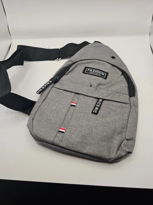 Grå crossbody-väska Fashion axelremsväska  - Snygg grå crossbody-väska i textil med svart justerbar axelrem. Väskan har flera fack med dragkedja, detaljer med texten 'NICE NIC' och små rödvita band. Praktisk metallring för hörlurar och stilren design. Perfekt för dig som vill ha en smidig och trendig väska.