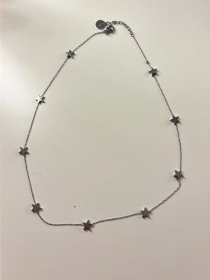 Silverfärgat halsband med stjärnor - Säljer ett tunt halsband i silverfärg med små stjärnformade hängen längs hela kedjan. Halsbandet har en enkel och stilren design och justerbar längd med förlängningskedja. Perfekt för dig som gillar minimalistiska smycken med en twist. Tyvärr sitter en av stjärnorna löst och det är en liten knut på kedjan men den syns inte när du har på dig halsbandet. 