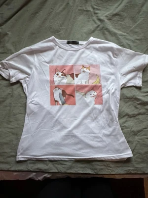 Vit t-shirt med kattmotiv - Vit t-shirt med fyrkantigt tryck av tecknade katter i olika poser på bröstet. Meme katter ;) T-shirten har korta ärmar och rund halsringning. Perfekt för dig som gillar gulliga och lekfulla motiv. Väldigt mjukt material. Köpt i storlek m från Kawaii.se men är liten i storlek och sitter mer som en s. Hör av er vid frågor!⭐️