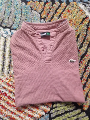 Rosa pikétröja från Lacoste - Säljer en rosa pikétröja från Lacoste med klassisk krage och knappar framtill. Tröjan har det ikoniska krokodilmärket på bröstet och är tillverkad i mjuk bomull. Perfekt för en avslappnad men ändå stilren look.