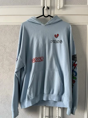 Ljusblå hoodie Polo G The Goat - Ljusblå hoodie med tryck från Polo G. Framsidan har texten 'POLO G', ett rött brustet hjärta och 'THE GOAT'. Färgglada motiv på ena ärmen. Klassisk känguruficka och huva. Perfekt för dig som gillar streetwear och unika prints.