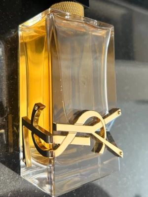 Yves Saint Laurent Libre parfym - Säljer en elegant parfym från Yves Saint Laurent, Libre. Flaskan är rektangulär i genomskinligt glas med guldiga detaljer och den ikoniska YSL-loggan i metall runt flaskan. Innehållet är bärnstensfärgat och korken har en lyxig guldstruktur. Se mängden på sista bild 😍