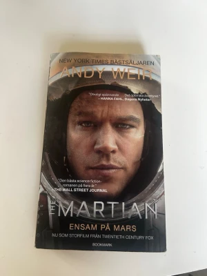 The Martian - Ensam på Mars - En spännande science fiction-roman av Andy Weir om astronauten Mark Watney som blir strandsatt på Mars. Boken är på svenska, har ett omslag i gråa och svarta toner med astronauttema och är utgiven av Bookmark. Perfekt för dig som gillar rymdäventyr och överlevnadsdrama.