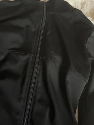 Svart sportjacka med dragkedja - Snygg svart sportjacka med hel dragkedja framtill och hög krage. Tillverkad i ett stretchigt och mjukt material som passar perfekt till träning eller löpning. Jackan har långa ärmar och en kroppsnära passform.