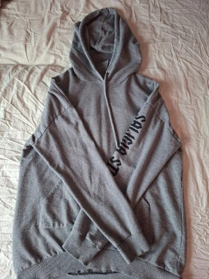 Grå hoodie med tryck på ärmen - Säljer en grå hoodie med huva och känguruficka. På ena ärmen finns ett svart tryck med texten 'SALCHA'. Hoodien har ribbade muddar vid ärmslut och nederkant. Perfekt för dig som gillar en enkel men snygg streetstil.