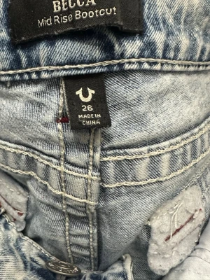 True Religion ljusblå bootcut jeans - Säljer ett par ljusblå bootcut jeans från True Religion, modell BECCA Mid Rise Bootcut. Jeansen har klassiska bakfickor med röd kontrastsöm och hål vid knäna