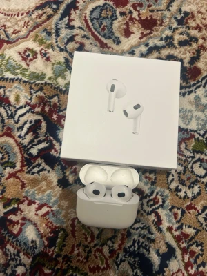 Apple AirPods 3  - Säljer ett par vita Apple AirPods 3 med trådlöst laddningsetui. Hörlurarna har en stilren och kompakt design i plast, med korta stjälkar och svarta detaljer. Levereras med originalkartong, laddningskabel och manualer.