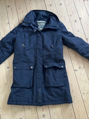 Marinblå Jacka från Sebago - Snygg marinblå jacka från Sebago med huva, stora fickor framtill och dragkedja med vindslå. Jackan har röda detaljer på insidan och är vadderad för extra värme. Perfekt för kyliga dagar och har en klassisk, tidlös design. Riktigt bra kvalitets jacka. Har använts ett fåtal gånger. Med slitage.