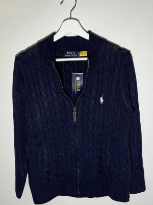 Mörkblå kabelstickad tröja Polo Ralph Lauren🤩🙌 - Mörkblå kabelstickad tröja från Polo Ralph Lauren med dragkedja framtill och ribbade muddar. Tröjan har det klassiska vita Polo-logotrycket på bröstet och två fickor framtill. Perfekt för en stilren och avslappnad look.🤩🙌