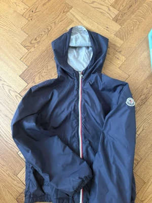 Mörkblå vindjacka från Moncler size 12A - Snygg mörkblå vindjacka från Moncler med huva och dragkedja med röd, vit och blå detalj. Jackan är storlek 1 och funkar för åldern 10-12 år. Jackan har ett Moncler-märke på ärmen och elastisk nederkant. Tillverkad i lätt, vindtätt material och har grått innerfoder. Nypris är 3995 :-
