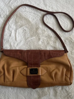 Brun axelväska från Tiger of Sweden - Snygg axelväska i två nyanser av brunt från Tiger of Sweden. Väskan har ett lock i mörkbrunt med krokopräglat mönster och en ljusbrun kropp i mjukt läder. Lång axelrem och metallskylt framtill med logga. Perfekt för dig som gillar klassisk men trendig stil.