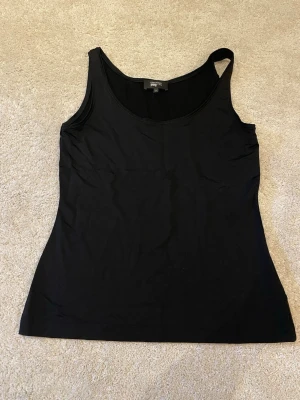 Svart linnetopp Essential Joyshop - Snygg svart linnetopp från Essential Item Joyshop. Toppen är ärmlös och har en enkel, stilren look som passar perfekt till jeans eller kjol. Tillverkad i 95% viskos och 5% elsatsningar för en  skön känsla hela dagen.