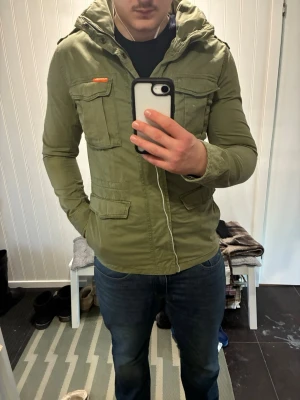 Fieldjacket grön från Superdry - Grön vindjacka med huva från Superdry. Jackan har fyra stora fickor framtill, patch med brittisk flagga på ärmen och stängs med dragkedja och tryckknappar. Snygga orange detaljer och normal passform.