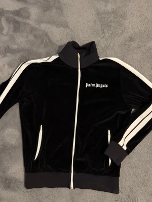 Svart Palm Angels ziptröja - Säljer en svart ziptröja från Palm Angels med vita ränder längs ärmarna och vit dragkedja. Tröjan har hög krage, muddar vid ärmslut och nederkant samt två fickor med dragkedja. Materialet är mjukt och har en lätt glansig finish.