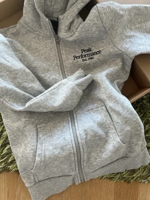 Grå hoodie från Peak Performance - Ljusgrå hoodie med dragkedja från Peak Performance. Har huva, två fickor framtill och broderad logga på bröstet med texten 'Peak Performance Est. 1986'. Något liten i storlek. 