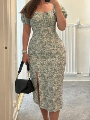 Gina Tricot grön blommig midi sommarklänning med slits - Supersöt midiklänning från Gina i grön och vit med blommigt mönster och puffärmar. Klänningen har en smickrande v-ringning, liten knytning framtill och en snygg slits på ena sidan. Perfekt för sommarens alla tillfällen! Använd 2, 3 gånger, jättefint skick. Storlek L. Klänninger har elastan.