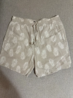 Beige shorts med palmblad från 157 - Säljer ett par beige shorts från 157 med vitt palmbladsmönster. De har elastisk midja med snörning och sidofickor. Perfekta för varma dagar och har en avslappnad passform. Materialet känns lätt och luftigt.